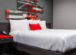 Modern hotel room at Astro Pasadena Hotel with a white bed, red accent pillow, gray walls, and contemporary artwork above the headboard.