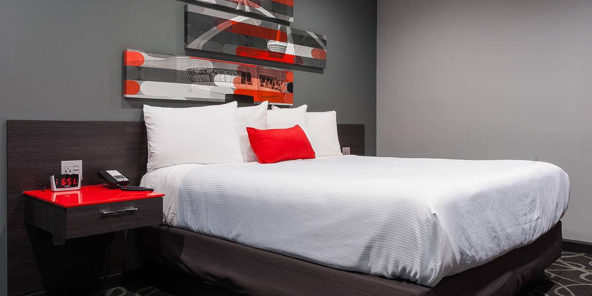 Modern hotel room at Astro Pasadena Hotel with a white bed, red accent pillow, gray walls, and contemporary artwork above the headboard.