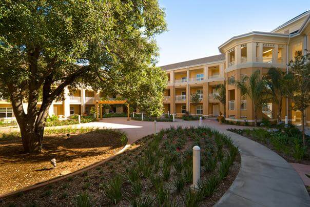 Serene Atherton Baptist Homes courtyard with winding path, greenery, and yellow building under blue sky