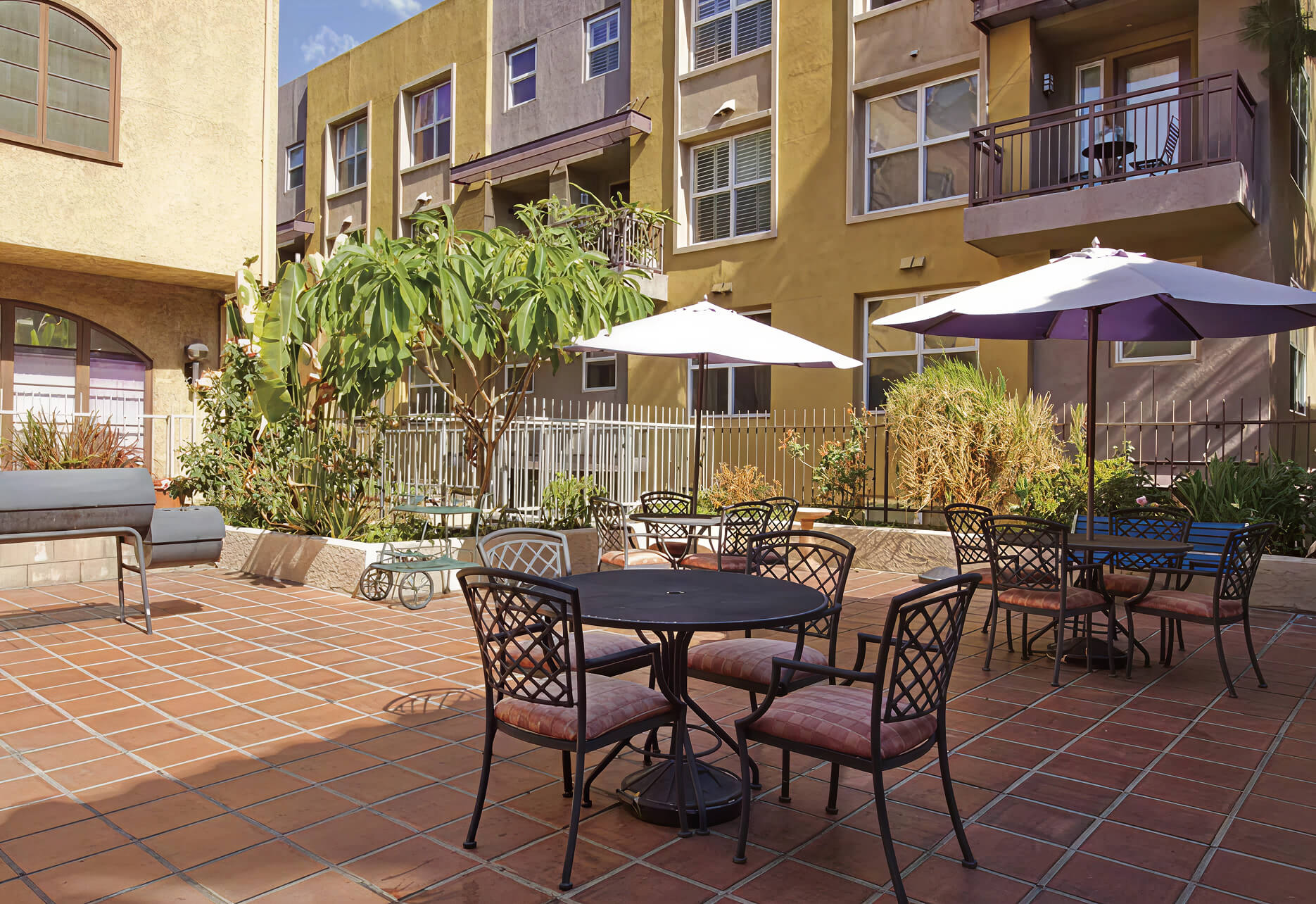 A sunny courtyard at Savant of Alhambra with tables, chairs, and umbrellas on a tiled patio.
