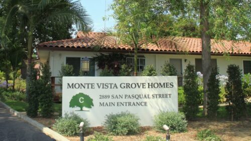 The main entrance sign for Monte Vista Grove Homes, showing the address and a tree logo amidst lush greenery.