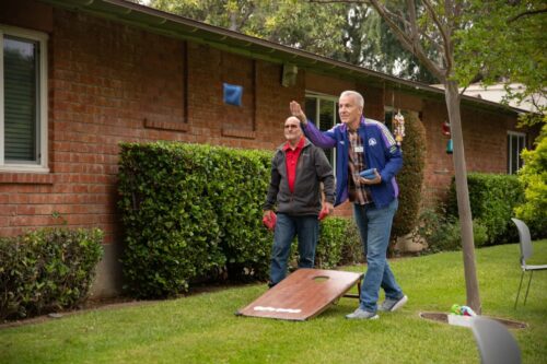 Two senior residents playing a casual game of cornhole (beanbag toss) on the grassy lawn outside a brick building