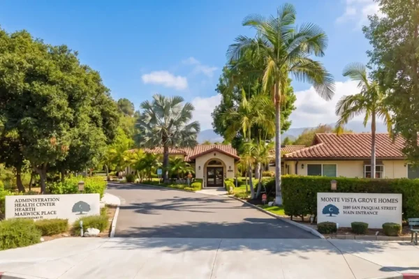 A symmetrical, bright daytime composition of a facility entrance. A paved driveway leads directly to an arched entryway of a terracotta-roofed, Spanish-style building, framed by a single central palm tree. Two large, identical white concrete signs are positioned on the left and right foreground. The left sign text reads: "ADMINISTRATION HEALTH CENTER HEARTH MARWICK PLACE" below a stylized tree logo. The right sign text reads: "MONTE VISTA GROVE HOMES 2889 SAN PASQUAL STREET MAIN ENTRANCE" below a stylized tree logo. To the right of the right sign is a green park bench and a lamppost. The foreground features a concrete sidewalk, with mountains in the background under a blue sky with light clouds.