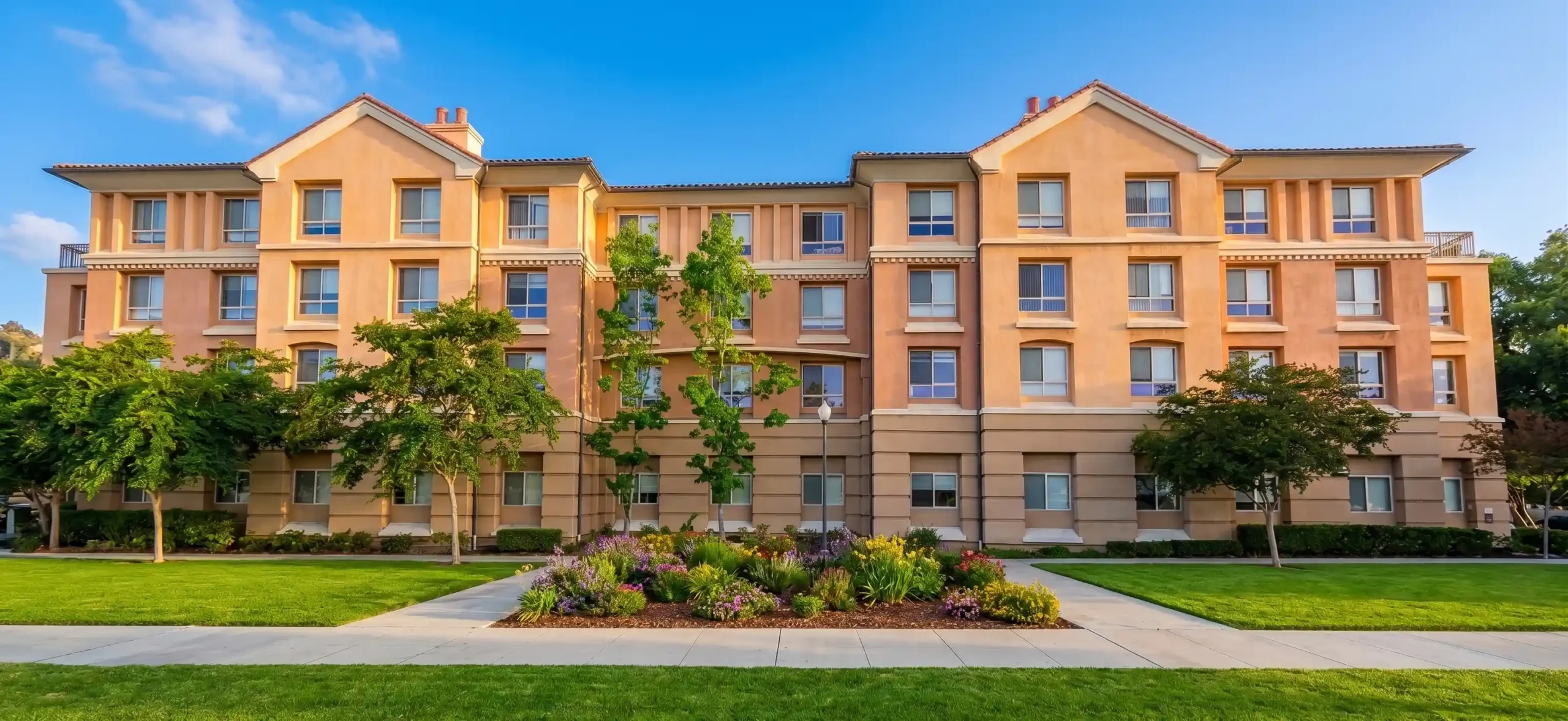 A wide-angle, symmetrical, and centered photograph of a large, modern, multi-story apartment complex. The facade, with its beige and light-brown stucco, two prominent gables, and multiple rows of windows, is bathed in warm, golden, late-afternoon sunlight. In the foreground, a precise concrete walkway leads from the bottom-center, dividing lush, vibrant green lawns, towards a central garden bed filled with colorful flowers in purple, yellow, and other multi-hued tones. Several well-manicured trees are positioned across the lawn and near the building's base. The perspective is direct and balanced, with rolling green hills visible in the far distance under a bright blue sky with light, wispy clouds. No text is clearly visible on the building.