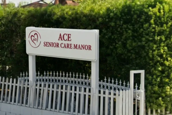 Entrance sign for ACE Senior Care Manor behind a white fence and dense green hedge in Pasadena, California.