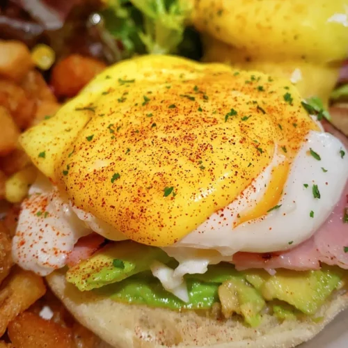 Close-up of eggs benedict with hollandaise sauce over a poached egg, avocado, and ham.