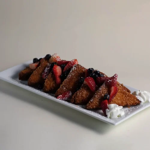 French toast sticks with strawberries, blueberries, powdered sugar, and whipped cream on a long white plate.