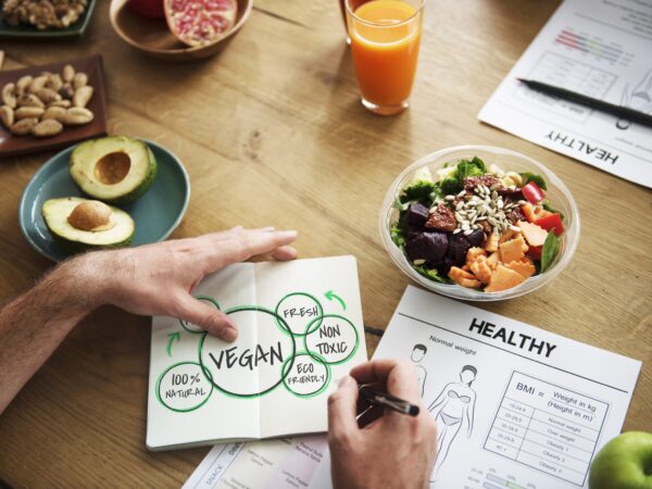 Healthy food flatlay featuring salad, avocado, and nuts; a person drawing a nutrition chart with vegan, fresh, and 100% natural labels, reflecting the True Food Kitchen philosophy.