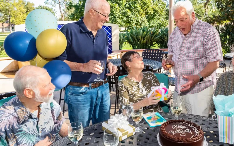 Group of older adults celebrating a birthday outdoors with balloons, cake, and gifts.