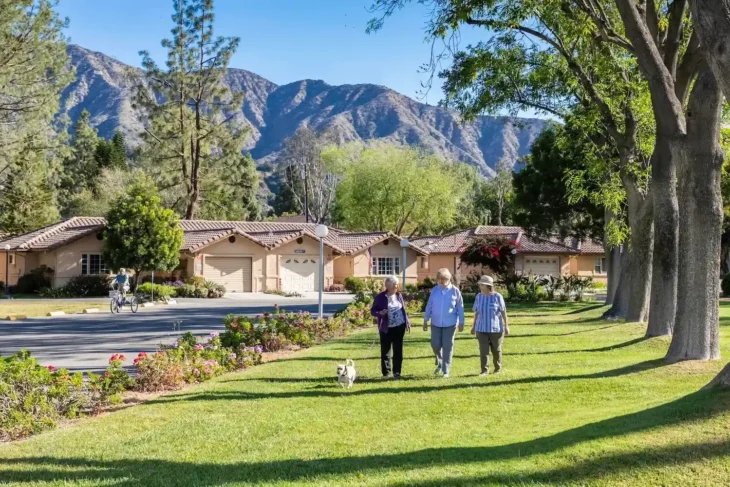 westminster-gardens-walking-lawn-mountain-view Older adults walking a dog along a landscaped green lawn with single-story cottages and mountain views in the background.