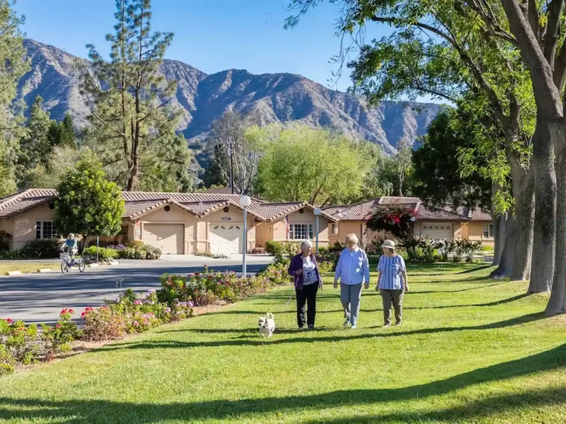Older adults walking a dog along a landscaped green lawn with single-story cottages and mountain views in the background.
