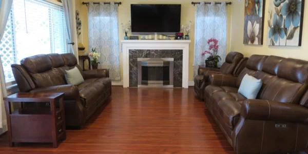 A bright and cozy communal living room with two large brown leather recliner sofas facing each other, a dark wood floor, and a white fireplace mantel with a large flat-screen TV mounted above it. Two large blue floral art pieces decorate the right wall.