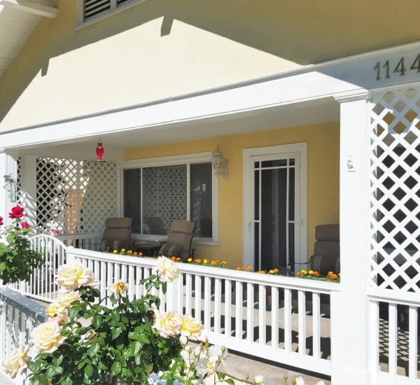The sunny exterior front porch of the El Molino Rose Villa house, showing the address "1144" and a white railing with trellis work. The porch is surrounded by blooming rose bushes in red, orange, and pale yellow.