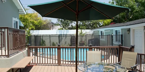 Outdoor wooden deck area with patio furniture and a large green umbrella, overlooking a secured swimming pool and spa area with a fence, set against the backdrop of the facility's exterior.
