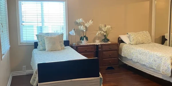 Semi-private resident room featuring two single adjustable hospital beds with patterned bedding, dark wood flooring, a ceiling fan, window blinds, and built-in closet space.