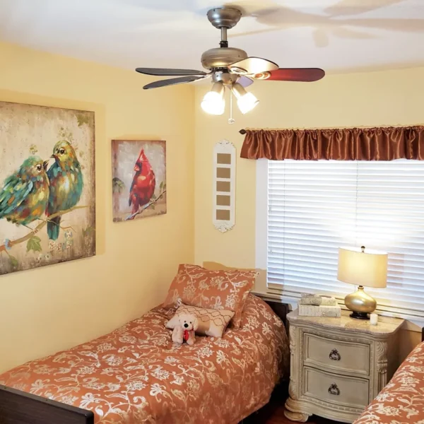 A bright double-occupancy bedroom with yellow walls, two twin beds with orange patterned bedding, a nightstand with a lamp between them, and a ceiling fan. The room is decorated with several large paintings of colorful birds.