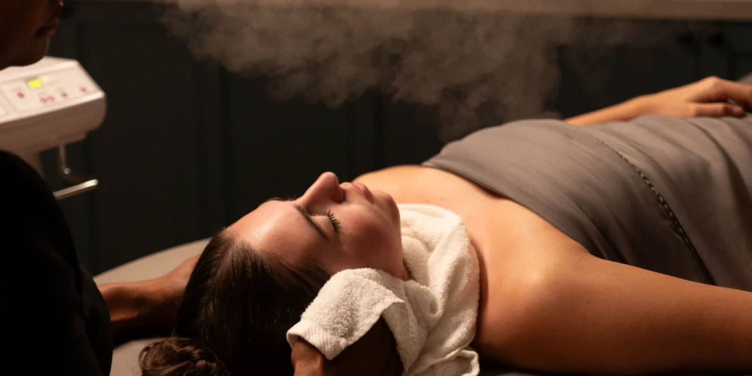Woman relaxing during a facial steam treatment at Burke Williams Day Spa, lying on a massage table with a warm towel under her neck.