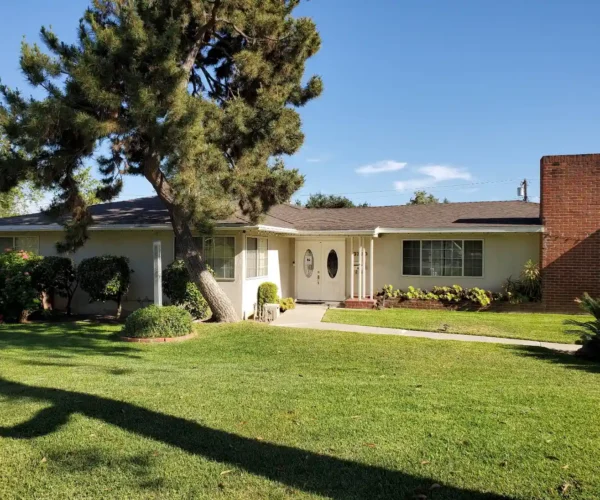 The exterior view of Hampton Guest Home in Pasadena, showing a welcoming single-story ranch-style house with a large pine tree, a manicured green lawn, a brick chimney, and white double entrance doors.