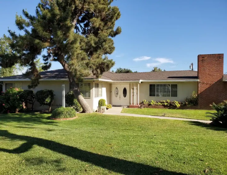 hampton-guest-home-exterior-building-3790-hampton-rd The exterior view of Hampton Guest Home in Pasadena, showing a welcoming single-story ranch-style house with a large pine tree, a manicured green lawn, a brick chimney, and white double entrance doors.