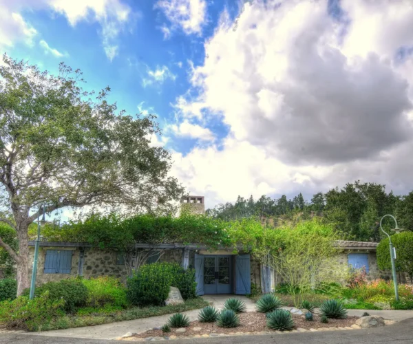 Front view of La Casita del Arroyo with stone walls, blue doors, desert plants, and a dramatic cloudy sky overhead.