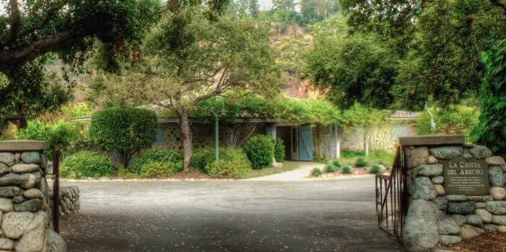 la-casita-del-arroyo-main-entrance-pasadena Historic stone building of La Casita del Arroyo surrounded by mature trees and lush greenery at the entrance in Pasadena.