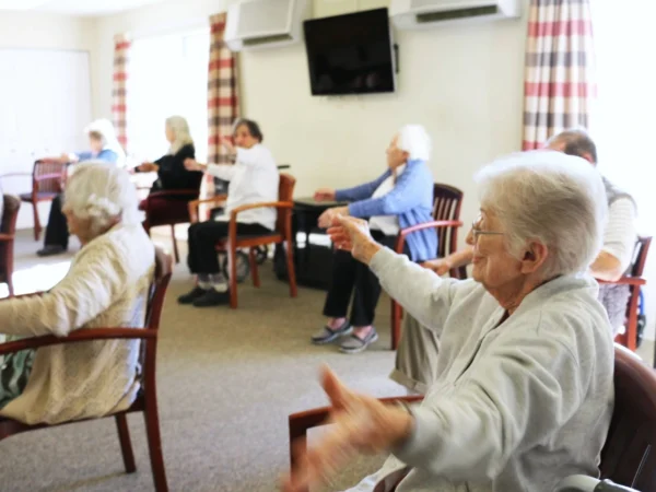 A group of senior residents participating in a seated exercise class in a common room at The Terraces at Park Marino, led by a smiling woman in the foreground.