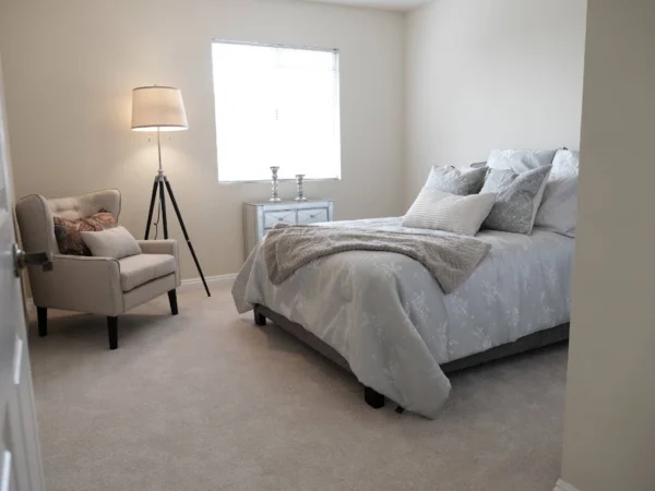 A brightly lit, modern senior living bedroom at The Terraces at Park Marino featuring a neatly made bed with grey and white linens, a comfortable beige armchair with a floor lamp, and a large window providing natural light.