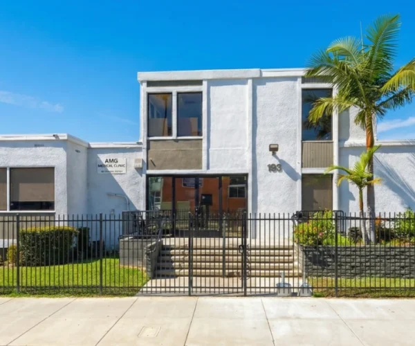 A wide-angle architectural shot of the AMG Medical Clinic at 193 E Orange Grove Blvd under a bright, cloudless blue sky. The composition is centered on the modern white and grey two-story medical building, framed by a lush palm tree on the right and a dark metal security fence in the foreground. Sharp midday sunlight highlights the textured stucco of the "AMG Medical Clinic" signage and the bold house number 193.