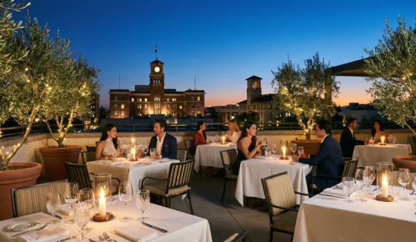 A wide, symmetrical shot of an elegant rooftop restaurant at twilight. The deep blue sky contrasts with the warm, flickering glow of candles on tables draped in white linen. Potted olive trees wrapped in delicate fairy lights frame the scene. In the distance, a historic clock tower is illuminated against the dusk, emphasizing a sophisticated, romantic atmosphere of urban fine dining.