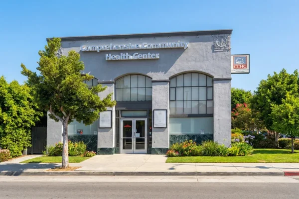 A bright, symmetrical architectural photograph of a one-story medical office under a clear, vibrant blue sky. The composition is centered and eye-level, highlighting the building's light grey facade and three large arched windows. Direct, midday sunlight illuminates the structure, casting soft shadows and making the large, silver 3D lettering that reads "Comprehensive Community Health Center" stand out on the upper facade. To the right, a square pylon sign displays the "CCHC" logo in orange and blue. The building is framed by lush green trees and manicured shrubbery, creating a professional and welcoming atmosphere.