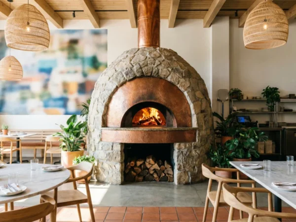 A wide, bright interior shot of a modern restaurant with high ceilings and exposed light wood beams. The focal point is a massive, dome-shaped stone oven with a copper chimney and a roaring fire inside. The room is filled with natural light, featuring white marble tables, light-wood chairs, lush green potted plants, and woven wicker pendant lights. A soft, abstract mural decorates the far wall.