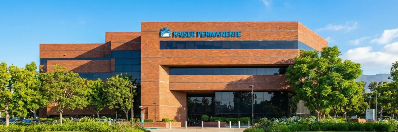 A bright, direct front photograph of the main entrance of the brick Kaiser Permanente building under a clear blue sky. A prominent "KAISER PERMANENTE" logo and text sign is centered on the upper facade above the windows. Sunlight creates distinct shadows, highlighting the textured brick and the lush, well-maintained landscaping with green lawn, bushes, and trees in the foreground. A paved driveway leads to the glass doors.