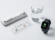 A minimalist, high-key medical flat lay on a clean, light gray surface. Bright, soft top-down lighting creates subtle, clean shadows. The composition features a white medical injection pen next to a transparent weekly pill organizer marked with "S M T W T F S" in black letters. A clear glass of water sits beside a black smartwatch, which displays the text "Health Goal Achieved" accompanied by a vibrant green checkmark icon.