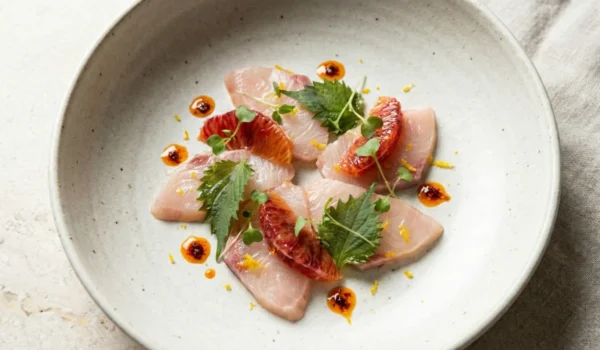 A minimalist top-down view of a Hamachi crudo dish served in a matte, speckled ceramic bowl. Translucent slices of fresh white fish are arranged symmetrically, garnished with bright blood orange segments, delicate microgreens, and small, precise droplets of reddish chili oil. The lighting is soft and even, highlighting the fresh textures against a neutral linen background.