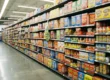 A low-angle, deep perspective shot of a long supermarket aisle receding toward a distant vanishing point. Warm overhead fluorescent lighting reflects off a polished gray floor, illuminating dense, symmetrical rows of colorful packaged goods and cereals that fill the frame from floor to ceiling.