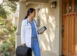A photorealistic medium shot of a female physician in white medical coat and blue scrubs standing on a sunlit residential porch. She carries a professional black medical bag and holds a digital tablet displaying patient charts. Bright afternoon sunlight casts soft diagonal shadows across the beige stucco wall. The text "DR. A. CHEN" is clearly embroidered in blue on the left side of the white coat.