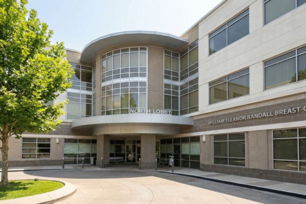 A wide-angle landscape photograph captures the sunlit exterior facade of a modern multi-story medical building under a clear blue sky. The architecture features a prominent curved glass and concrete entrance section labeled "NORTH LOBBY" in white capital letters above a canopy. To the right, on a textured concrete wall, the text "WILLIAM ELEANOR RANDALL BREAST CENTER" is clearly visible. A paved circular driveway leads to the covered entrance, and a large green tree with dense foliage stands on a grassy island to the left, casting shadows. The scene is brightly lit by direct sunlight, highlighting the textures of the concrete, glass windows, and asphalt.