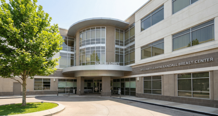A wide-angle landscape photograph captures the sunlit exterior facade of a modern multi-story medical building under a clear blue sky. The architecture features a prominent curved glass and concrete entrance section labeled "NORTH LOBBY" in white capital letters above a canopy. To the right, on a textured concrete wall, the text "WILLIAM ELEANOR RANDALL BREAST CENTER" is clearly visible. A paved circular driveway leads to the covered entrance, and a large green tree with dense foliage stands on a grassy island to the left, casting shadows. The scene is brightly lit by direct sunlight, highlighting the textures of the concrete, glass windows, and asphalt.