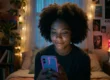A medium shot captures a young Black woman with voluminous natural hair smiling gently, her face illuminated by the cool blue light emanating from the smartphone she holds in her hands. She is in a cozy, dimly lit bedroom, filled with warm ambient light from a pink lava lamp and a strand of string lights on the wall. Posters (one partially reads "KALEWIK RIVIR"), books on a shelf, and plants create a bohemian feel. The composition is intimate, focusing on her interaction with the phone.