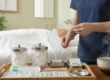 A medium close-up shot focused on a wooden treatment tray in a clean clinical environment. The composition highlights the hands of a practitioner in blue scrubs holding a cluster of colorful capped needles in the upper right. On the tray are organized glass jars of cotton balls, sealed metal supply packs, and a steaming ceramic bowl in the foreground. The background features a blurred patient resting on a white table and an unreadable soft-focus diagram on the pale green wall under diffused natural light.