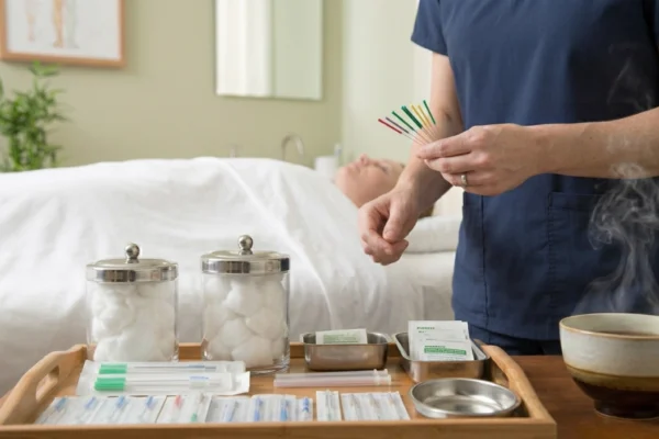 A medium close-up shot focused on a wooden treatment tray in a clean clinical environment. The composition highlights the hands of a practitioner in blue scrubs holding a cluster of colorful capped needles in the upper right. On the tray are organized glass jars of cotton balls, sealed metal supply packs, and a steaming ceramic bowl in the foreground. The background features a blurred patient resting on a white table and an unreadable soft-focus diagram on the pale green wall under diffused natural light.