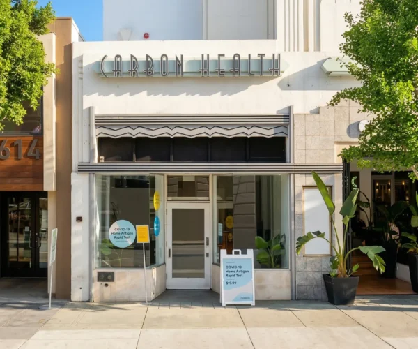 A symmetrical, eye-level exterior shot of the Carbon Health clinic facade under a clear blue sky. The words "CARBON HEALTH" are displayed in prominent, stylized silver metallic lettering above a glass entrance. The scene is lit by direct, bright daylight. In the windows, white and blue signs read "COVID-19 Home Antigen Rapid Test $19.99." Green trees flank the clean concrete sidewalk in the foreground.