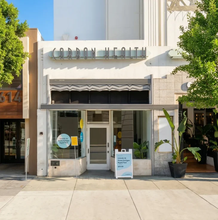 carbon-health-urgent-care-los-angeles-entrance A symmetrical, eye-level exterior shot of the Carbon Health clinic facade under a clear blue sky. The words "CARBON HEALTH" are displayed in prominent, stylized silver metallic lettering above a glass entrance. The scene is lit by direct, bright daylight. In the windows, white and blue signs read "COVID-19 Home Antigen Rapid Test $19.99." Green trees flank the clean concrete sidewalk in the foreground.