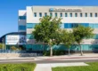A brightly lit, direct-angle photograph of the modern, four-story concrete and glass facade of the City of Hope, South Pasadena medical facility under a clear blue sky. Two mature leafy trees frame the center, casting distinct shadows on the clean sidewalk and pristine lawn. Clear text at the top center reads: 'City of Hope' with a logo, and 'SOUTH PASADENA'. On the far left, a curving canopy shelters a large banner-mural that proudly reads: 'Proud to rank among the nation’s top 10 ‘Best Hospitals’ for cancer', surrounded by small portraits. Blue railings line the access ramp and stairs.