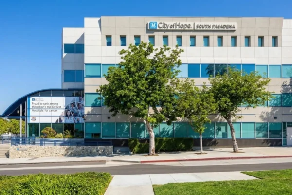 A brightly lit, direct-angle photograph of the modern, four-story concrete and glass facade of the City of Hope, South Pasadena medical facility under a clear blue sky. Two mature leafy trees frame the center, casting distinct shadows on the clean sidewalk and pristine lawn. Clear text at the top center reads: 'City of Hope' with a logo, and 'SOUTH PASADENA'. On the far left, a curving canopy shelters a large banner-mural that proudly reads: 'Proud to rank among the nation’s top 10 ‘Best Hospitals’ for cancer', surrounded by small portraits. Blue railings line the access ramp and stairs.