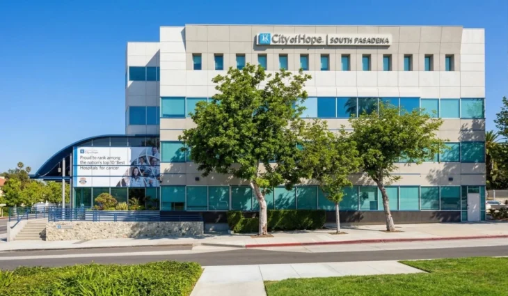 city-of-hope-south-pasadena-hospital-facade-supportive-care-in-cancer A brightly lit, direct-angle photograph of the modern, four-story concrete and glass facade of the City of Hope, South Pasadena medical facility under a clear blue sky. Two mature leafy trees frame the center, casting distinct shadows on the clean sidewalk and pristine lawn. Clear text at the top center reads: 'City of Hope' with a logo, and 'SOUTH PASADENA'. On the far left, a curving canopy shelters a large banner-mural that proudly reads: 'Proud to rank among the nation’s top 10 ‘Best Hospitals’ for cancer', surrounded by small portraits. Blue railings line the access ramp and stairs.