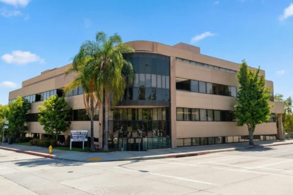 A wide-angle shot of a tan, three-story medical building with a prominent curved glass corner, captured in soft afternoon light. Lush green trees and palm trees frame the building against a clear sky. A sign on the corner reads "MEDICAL SPACE IMMEDIATELY FOR LEASE." The composition shows a clean, professional street-side view.
