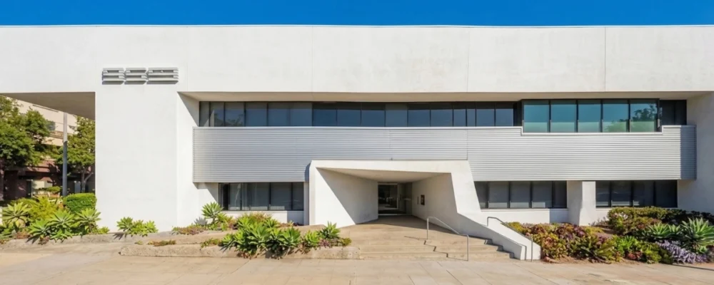 Wide-angle, symmetrical photograph of a stark white, modernist medical building facade under a clear blue sky and bright daylight. The ground-level entrance is framed by a distinctive, angular concrete step-up archway. The building features a long strip of second-floor windows screened by horizontal metallic louvers. On the far left, a textured, stylized numerical '999' is mounted on the wall. A lush, foreground garden of various succulents sits at the foot of the building along a sidewalk.