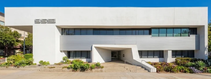 modern-pasadena-orthopedic-building-facade Wide-angle, symmetrical photograph of a stark white, modernist medical building facade under a clear blue sky and bright daylight. The ground-level entrance is framed by a distinctive, angular concrete step-up archway. The building features a long strip of second-floor windows screened by horizontal metallic louvers. On the far left, a textured, stylized numerical '999' is mounted on the wall. A lush, foreground garden of various succulents sits at the foot of the building along a sidewalk.
