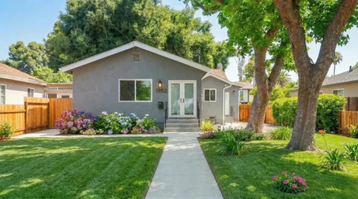 modern-pension-exterior-at-home-dr-visit-service A professional, wide-angle real estate photograph of a modern gray one-story house. The composition is perfectly symmetrical, with a clean concrete path leading directly to a white double door. The scene is bathed in bright, warm natural sunlight, creating soft shadows from a large leafy tree on the right. The foreground features a lush, vibrant green lawn and small clusters of blue and pink hydrangeas near the building. The architecture is simple and clean, with the number "1952" clearly visible in black font on the gray wall next to the entrance. The overall mood is peaceful, welcoming, and high-end.